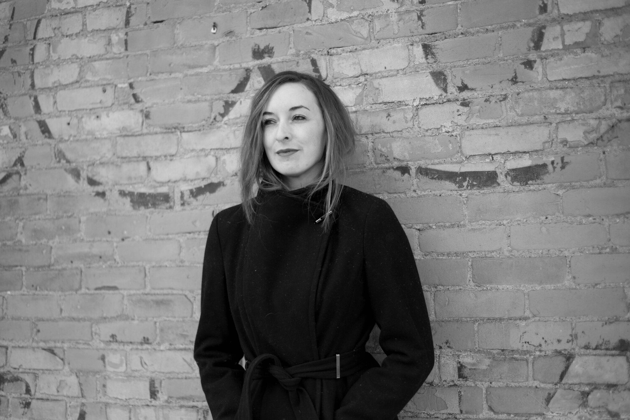 black and white photograph of poet Mairead Small Staid in a black coat standing in front of a brick wall. Photo by Jenna Meacham