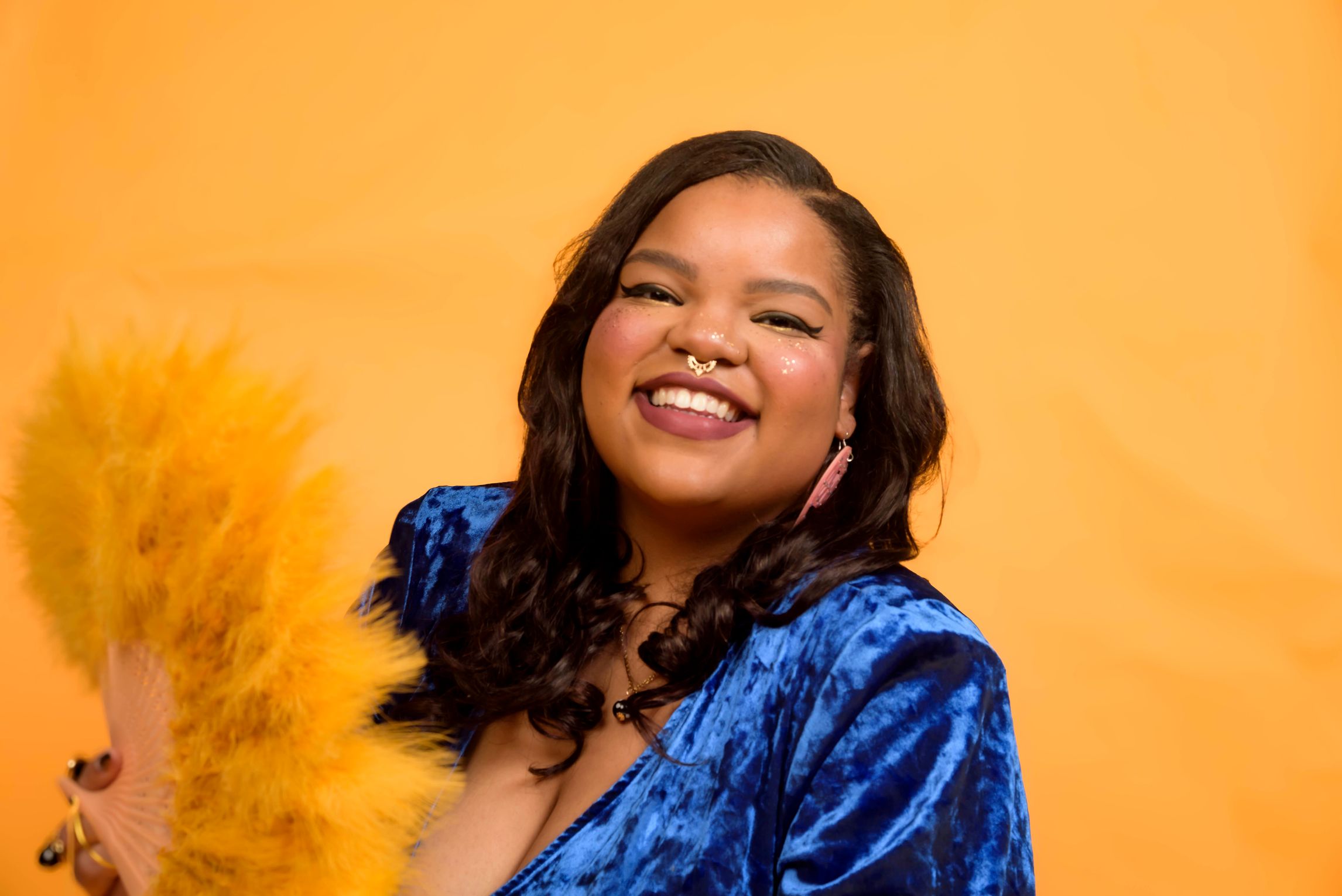 Headshot of m. mick powell, a Black femme person with a septum ring smiling and wearing a velvet blue blouse against a gold backdrop
