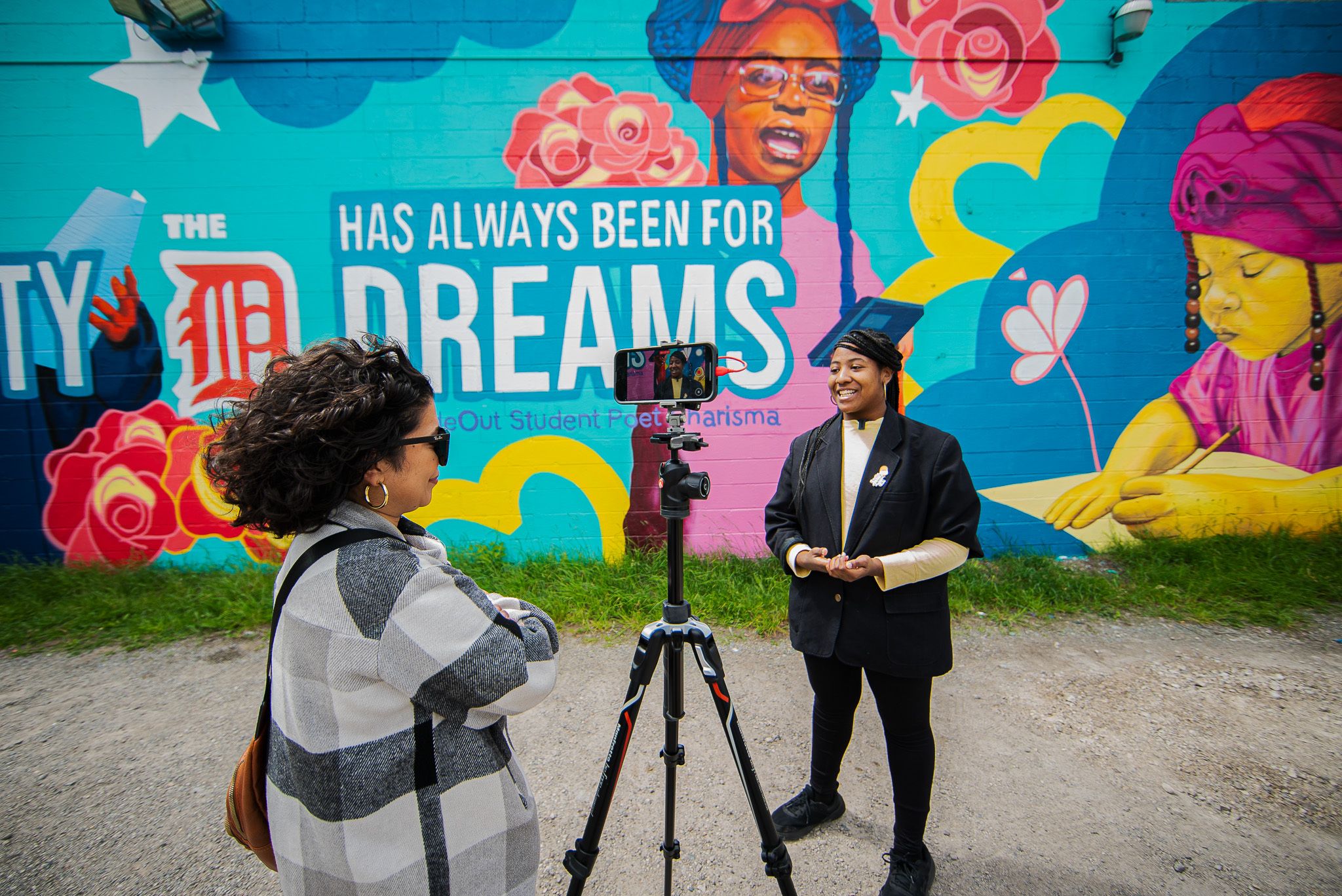 Someone behind and in front of a camera on a tripod. The backdrop is a colorful outdoor mural with text The D Has Always Been for Dreams.