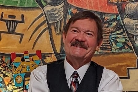 A man with a mustache wearing a suit vest smiling in front of a mural