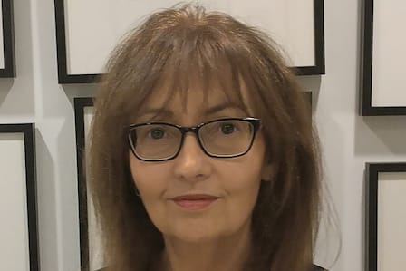 Woman with brunette hair and glasses smiling in front of a wall of picture frames