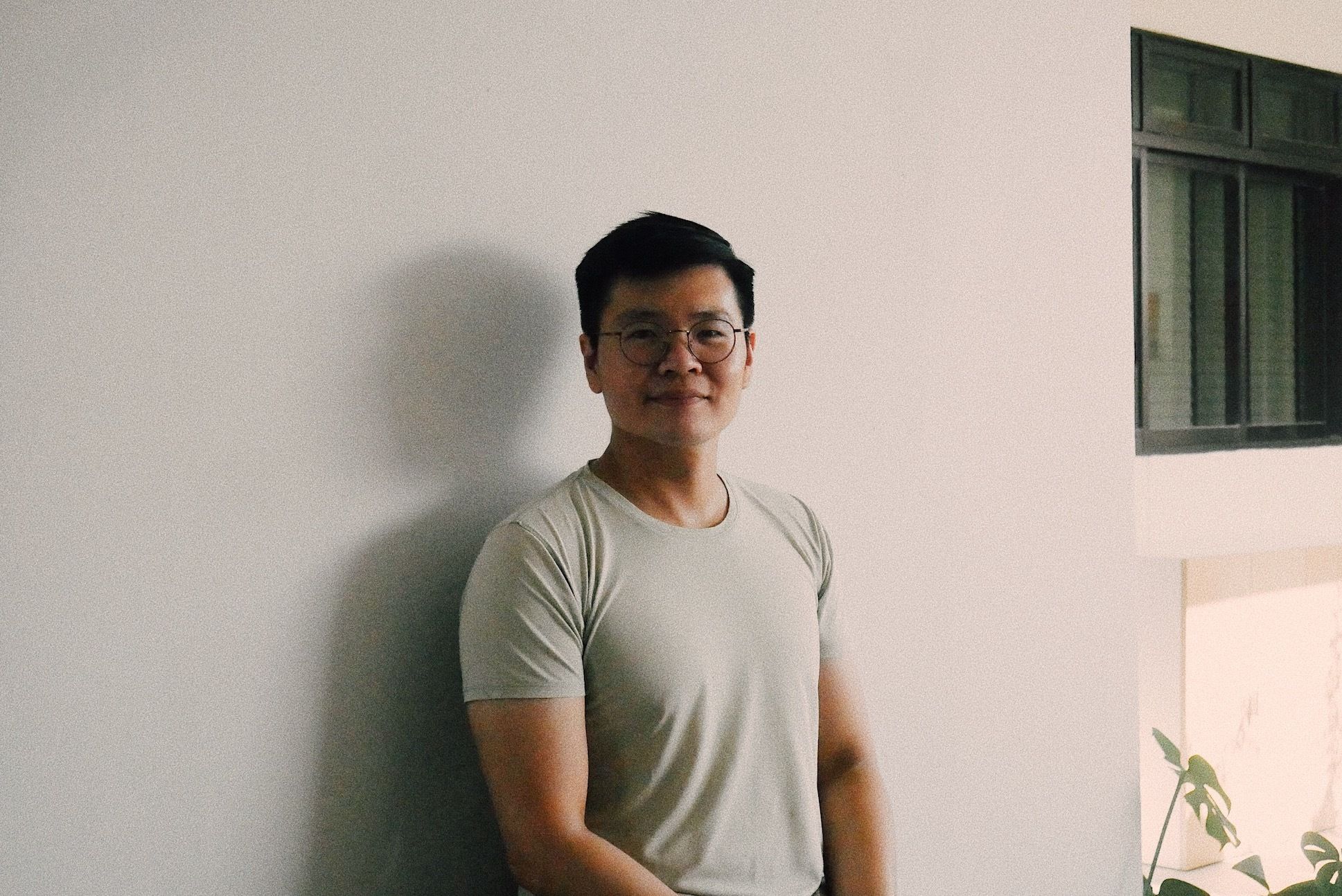 An Asian man with glasses and a white t-shirt standing in front of a white wall.