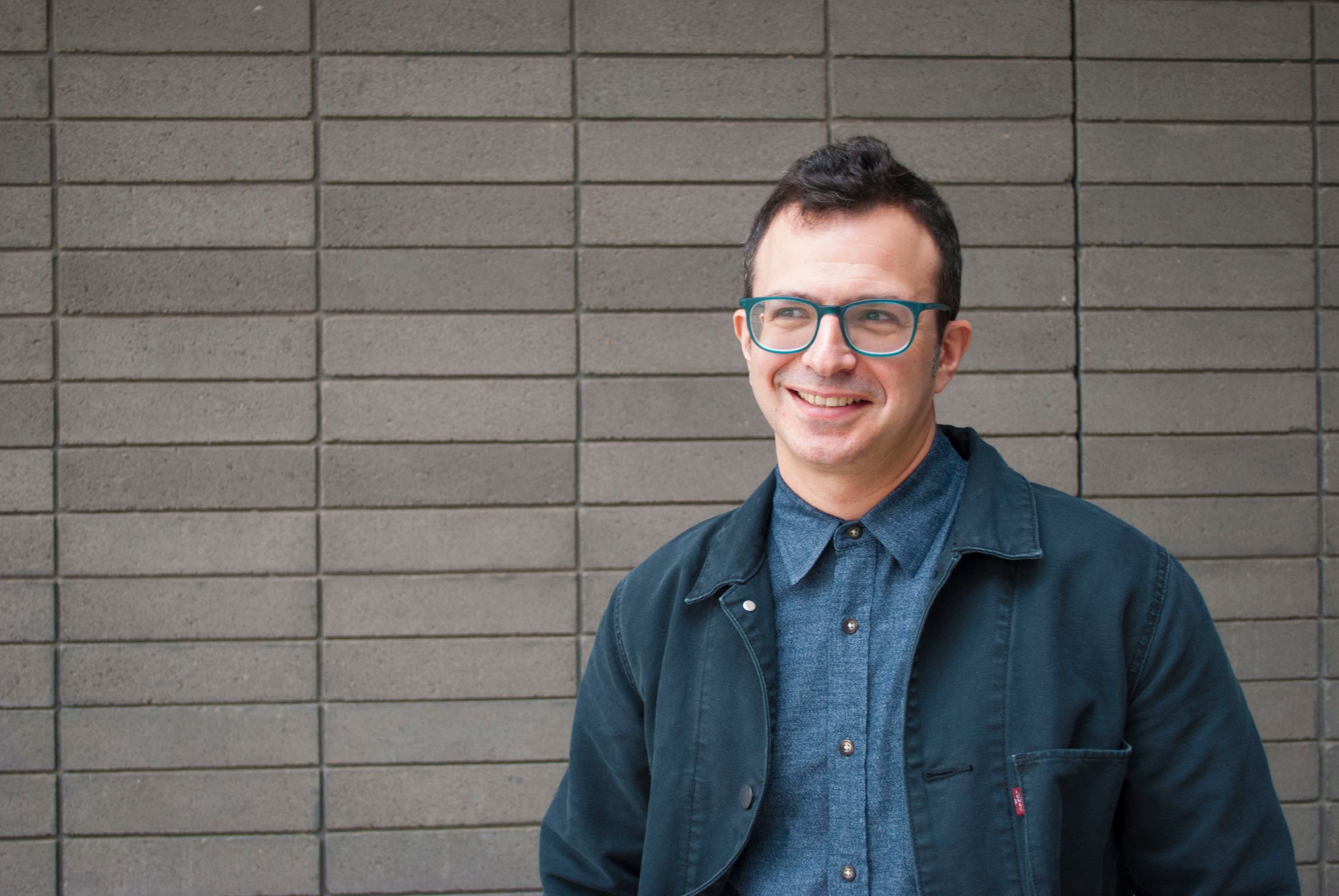 A man with brown hair and blue glasses in front of a brick wall