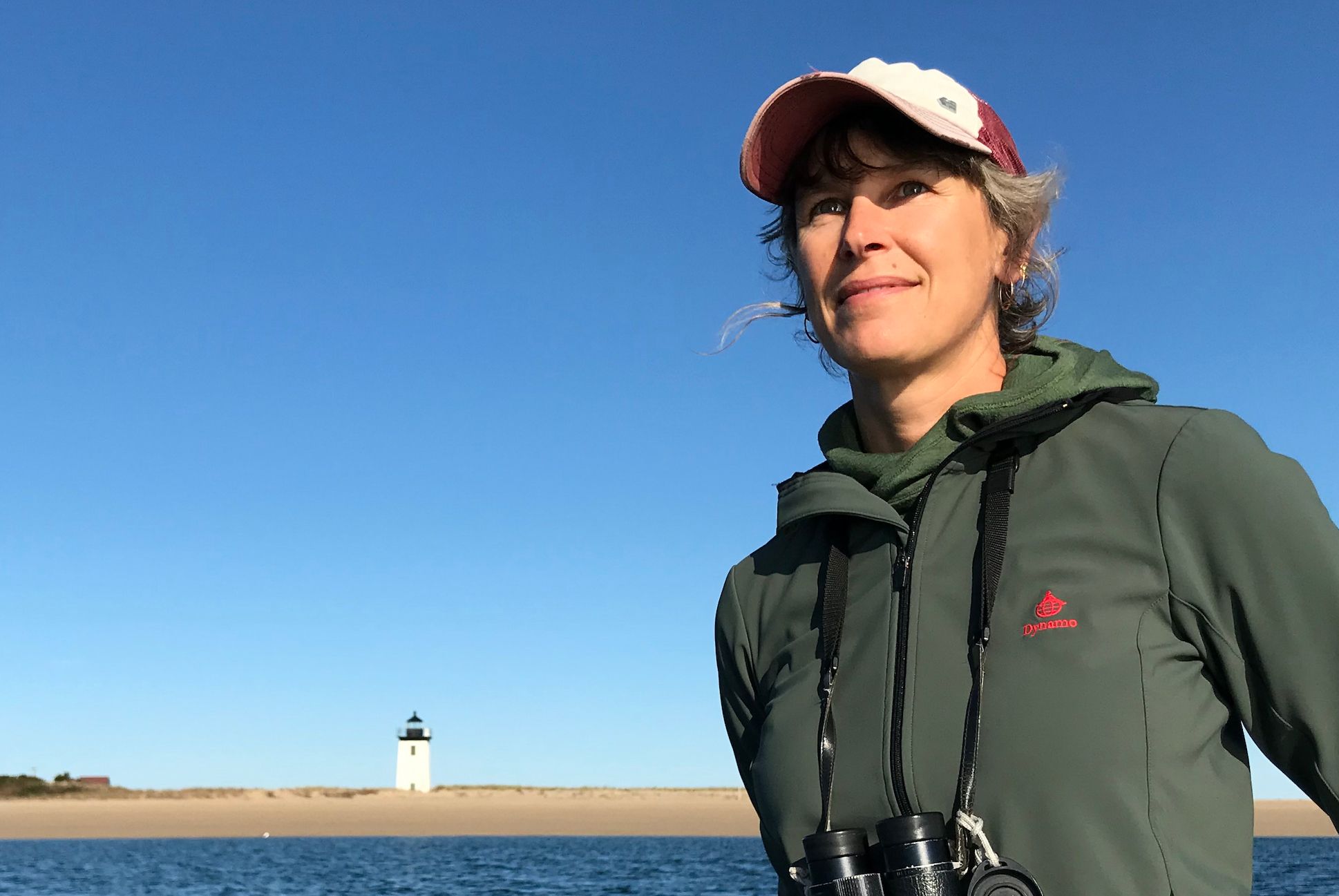 A white woman in a cap and binoculars standing by the water with a lighthouse in a far background