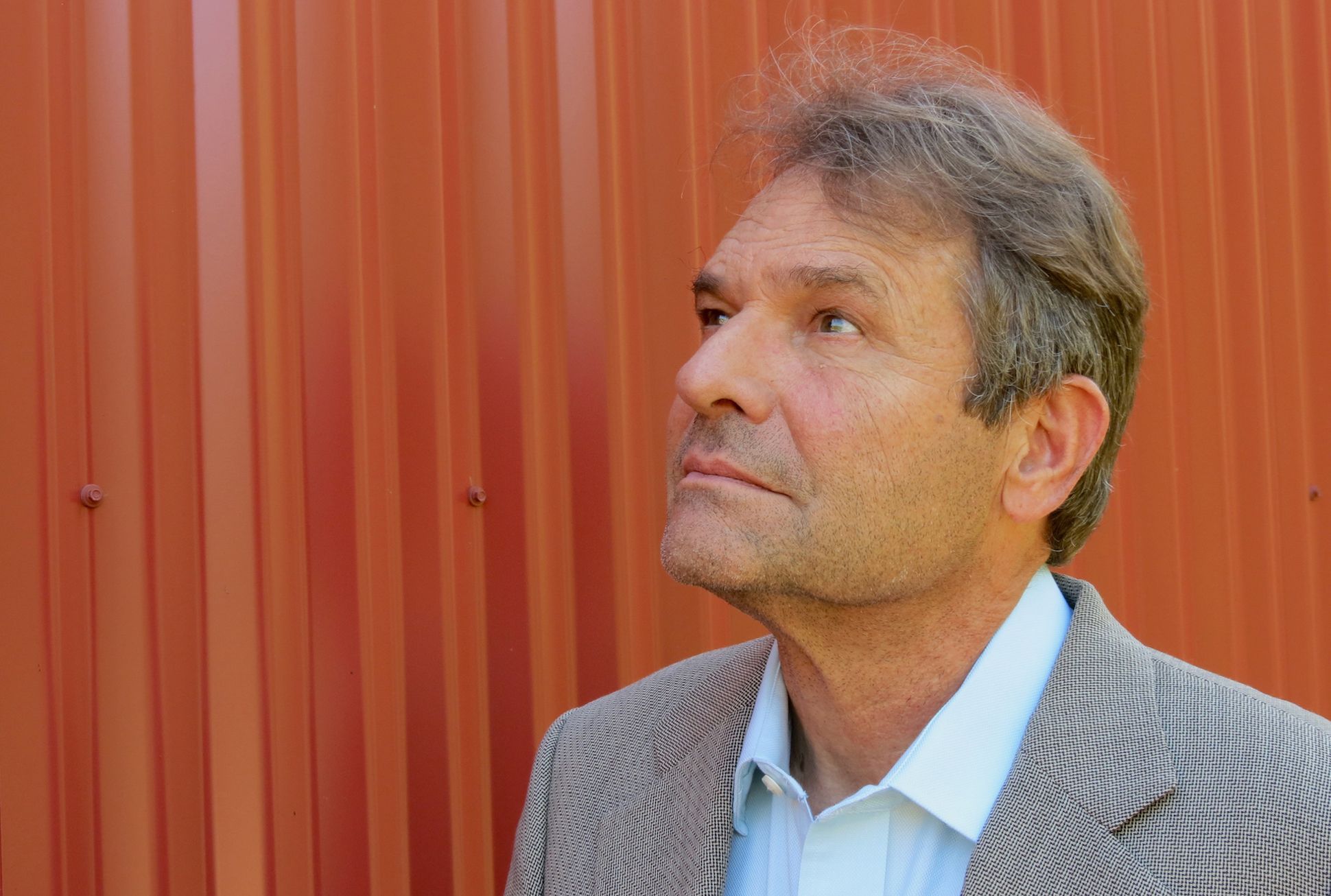 The writer Denis Johnson in a suit, posing in front of a bright orange wall.