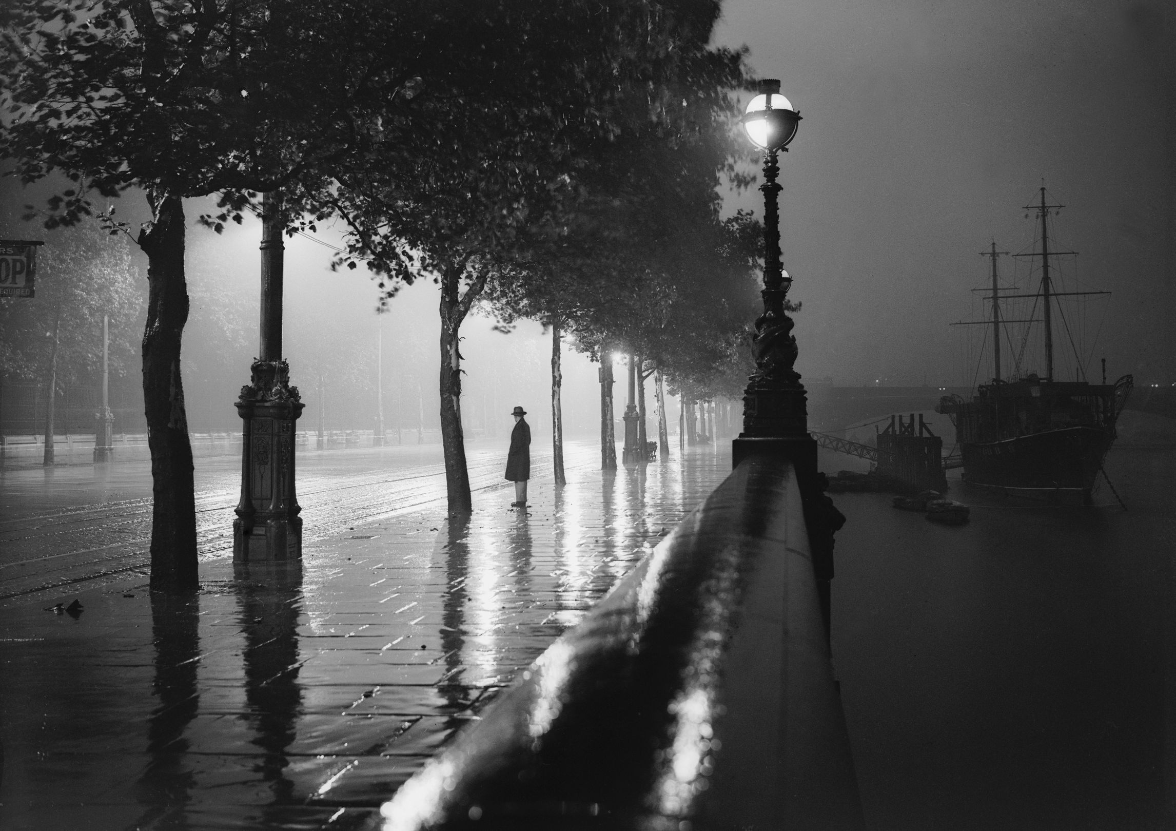 A man standing alone on a rain-drenched pavement on the River Thames Embankment, London.