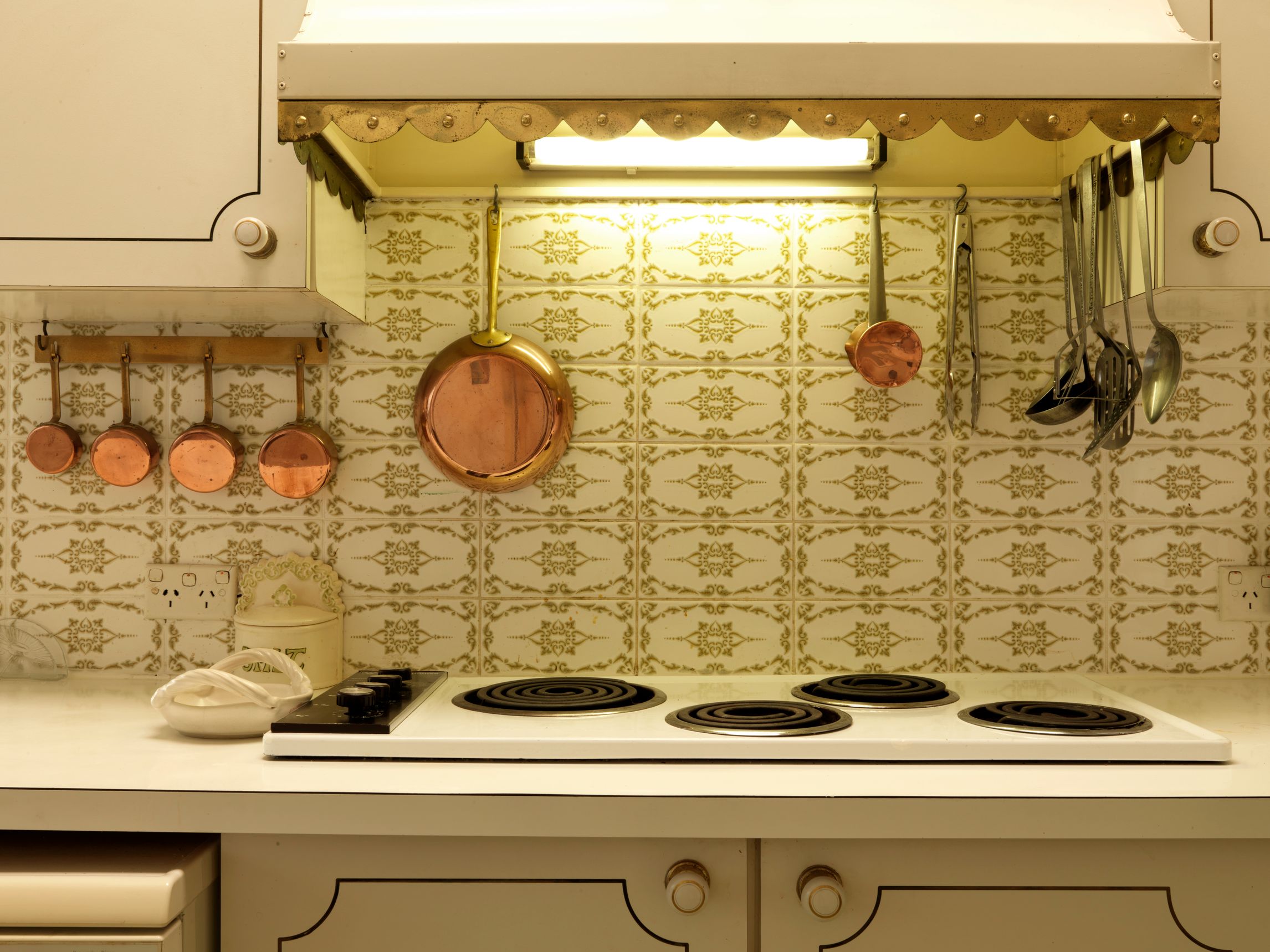 Portion of a retro kitchen, view of brass pots and utensils against white-and-green patterned wallpaper, an electric stovetop with an exhaust hood above with bright fluorescent light. cream colored counters.