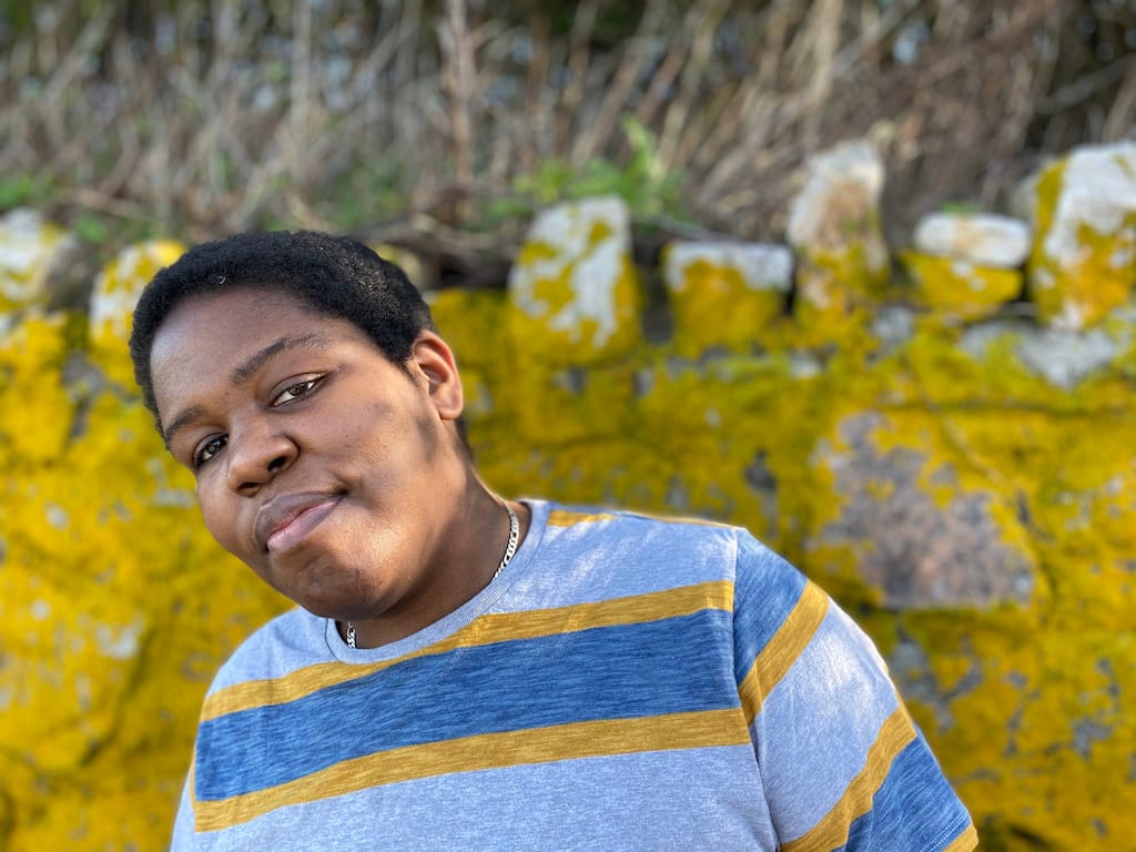 Candace Williams, a Black queer person with short black hair, stands with a mossy wall in the
background