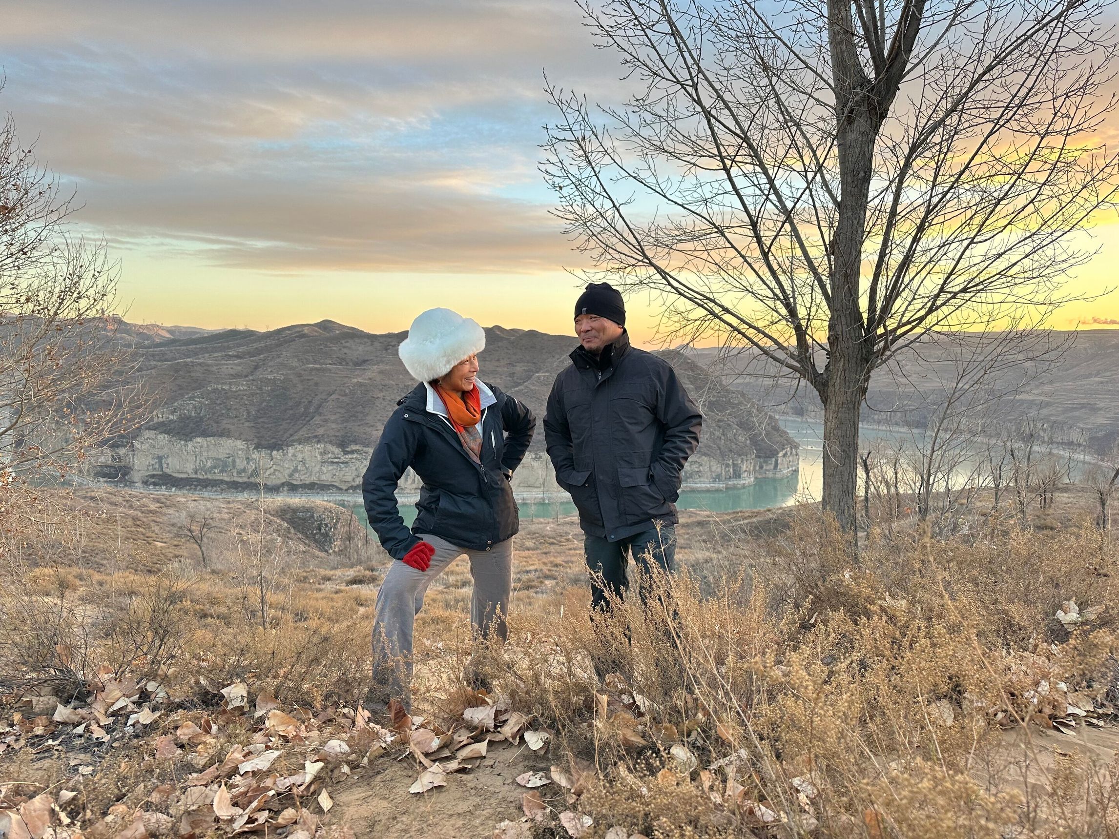 Two people wearing hats and coats pose in front of a river and rock formations