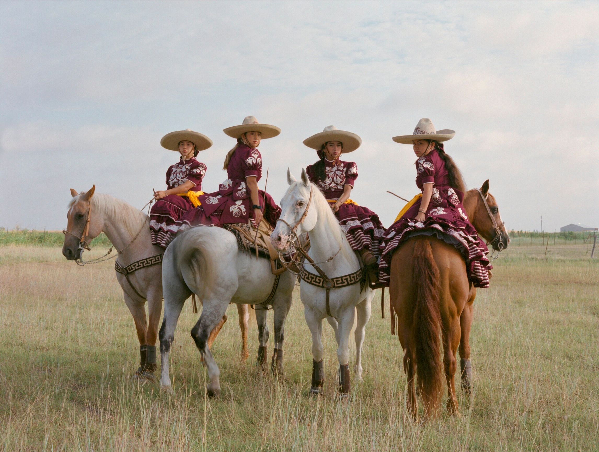 Four women in maroon and white traditional Mexican dresses and hats sit sidesaddle on horses. Two horses face the camera and two face away.