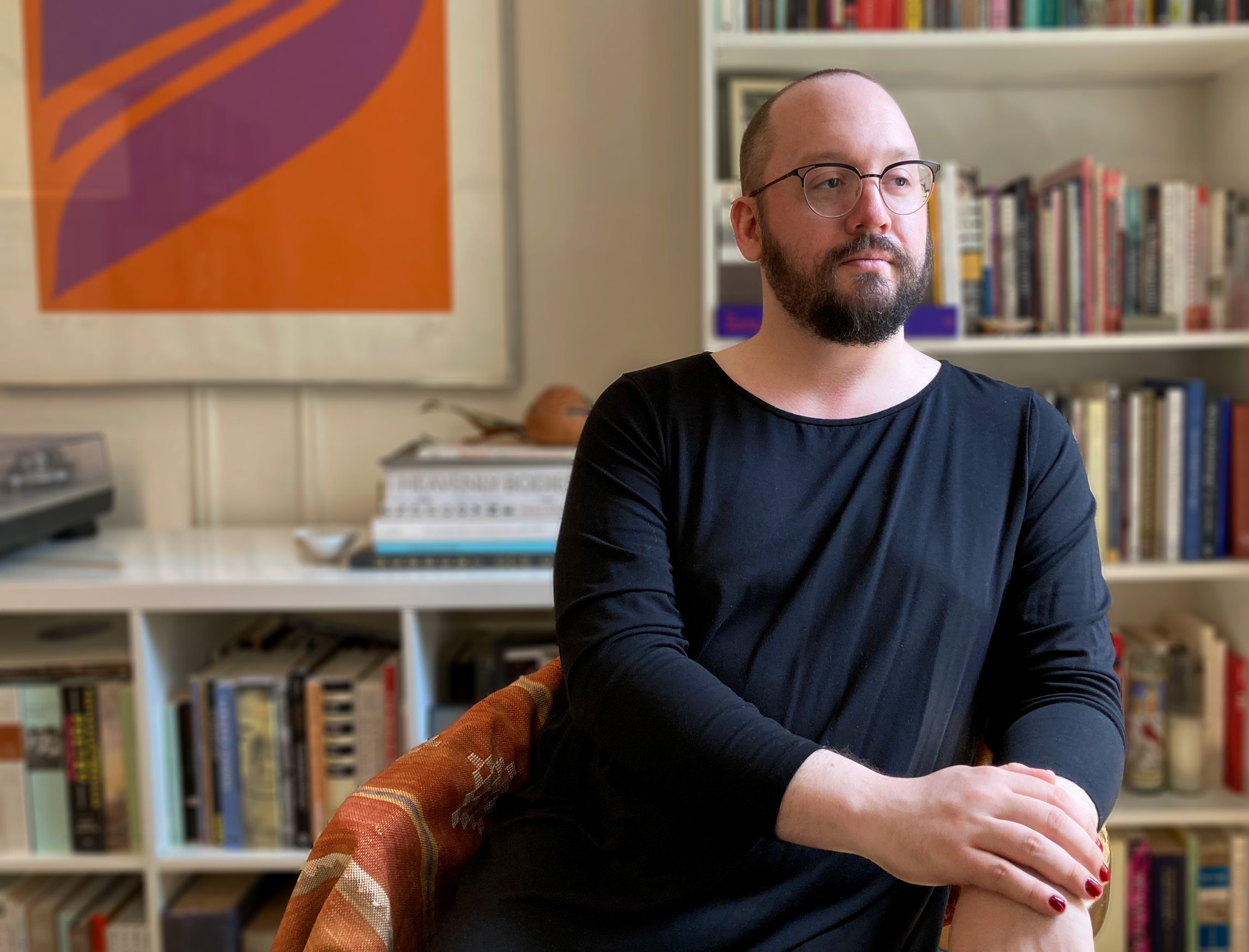 Photo of Trevor Ketner seated in front of a bookshelf. They are wearing a long black top, round glasses, and red nail polish.