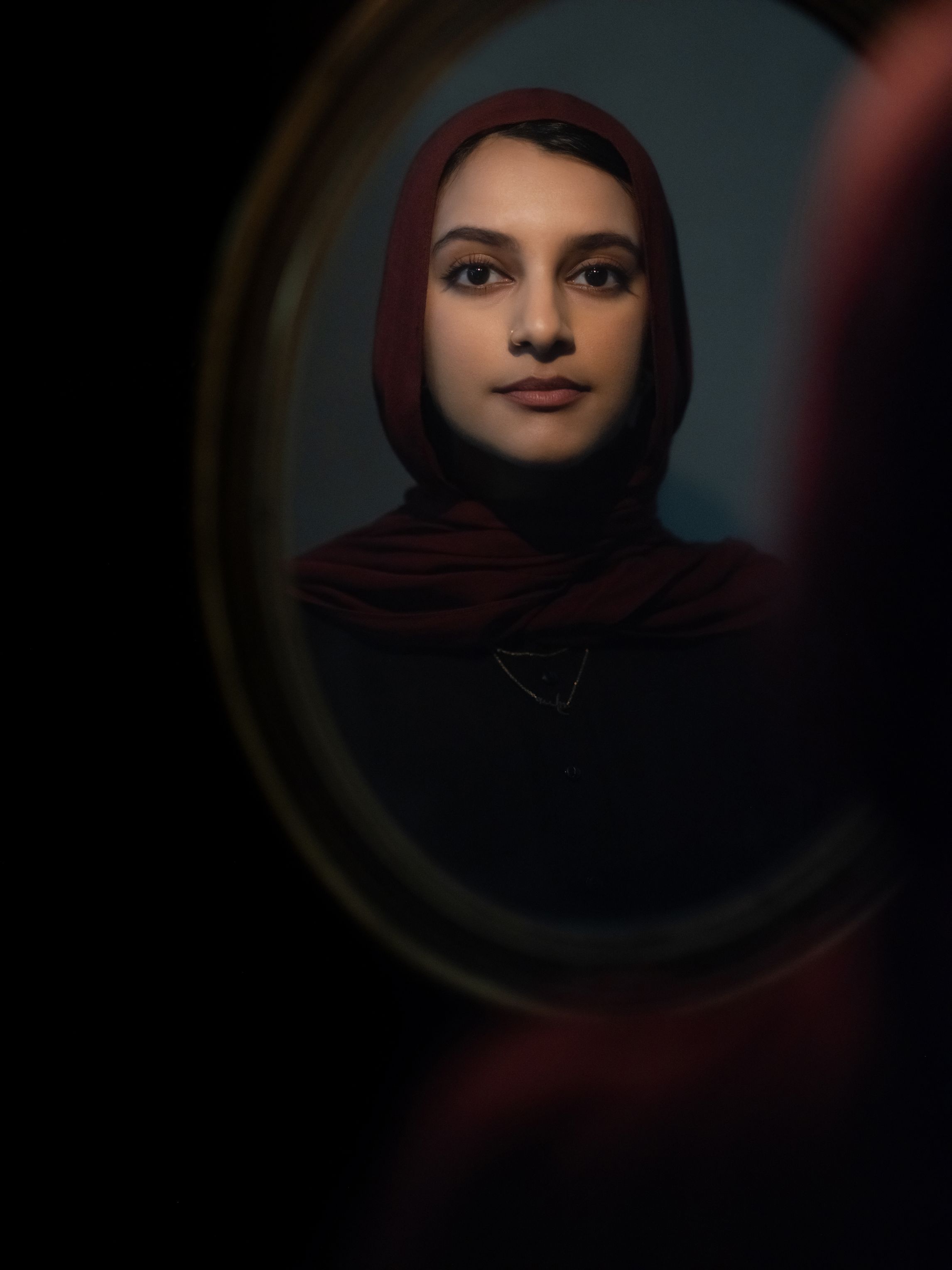 Sarah Ghazal Ali, a Pakistani woman in a maroon scarf, looks into a mirror.