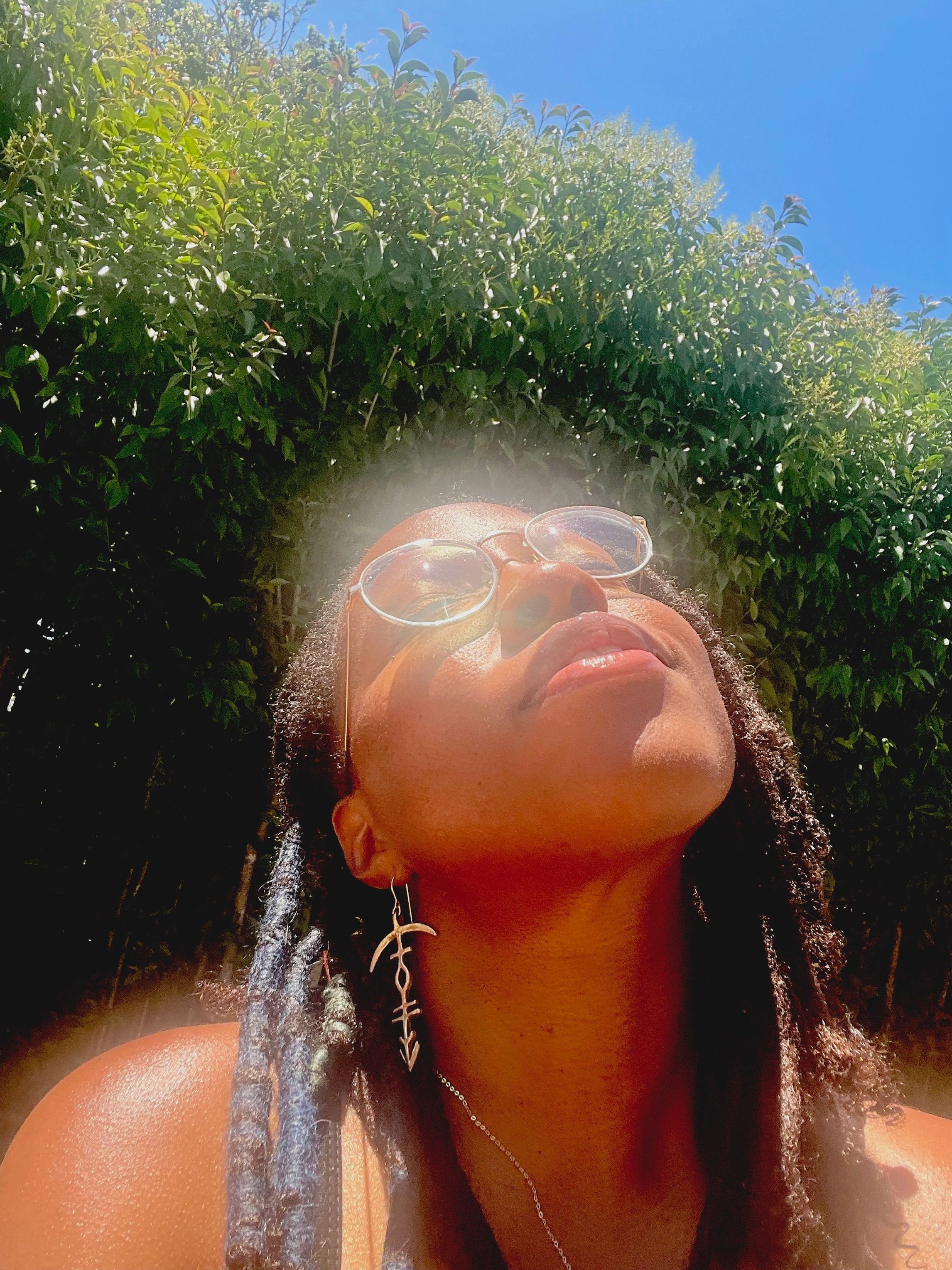 fahima ife, a Black woman with dark brown dreadlocks and gold rimmed glasses, sits outside with her face upturned towards the sun.