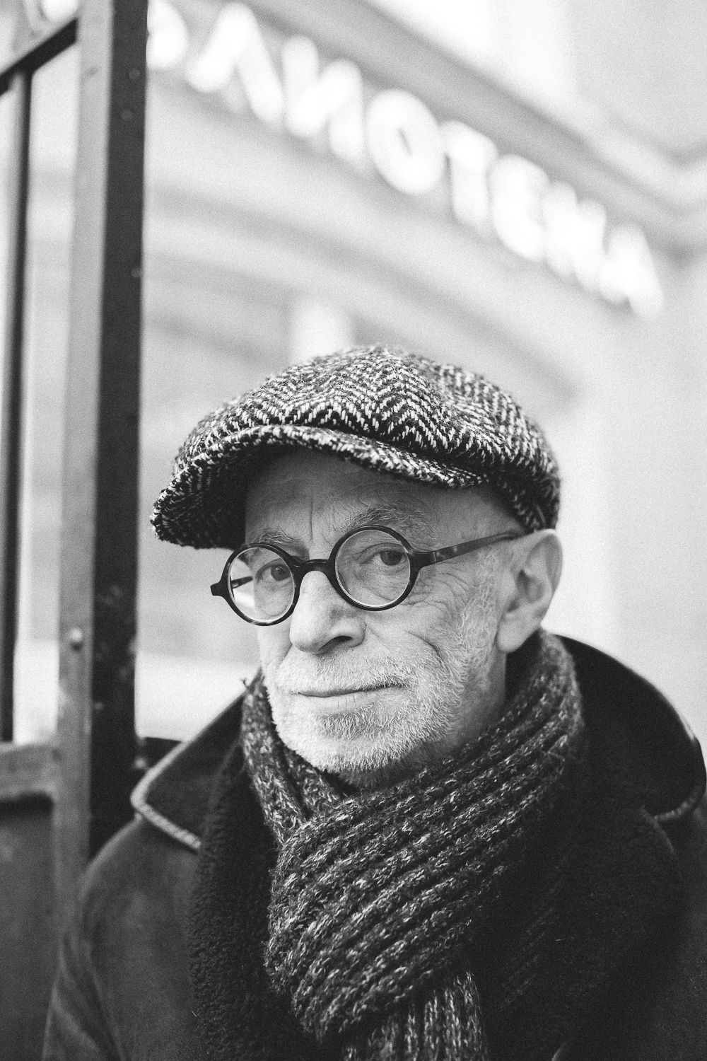 Black and white photo of Lev Rubinstein outdoors, a building with Russian letters is blurred in the background. Rubinstein is wearing glasses, a flat cap, and a thick scarf.
